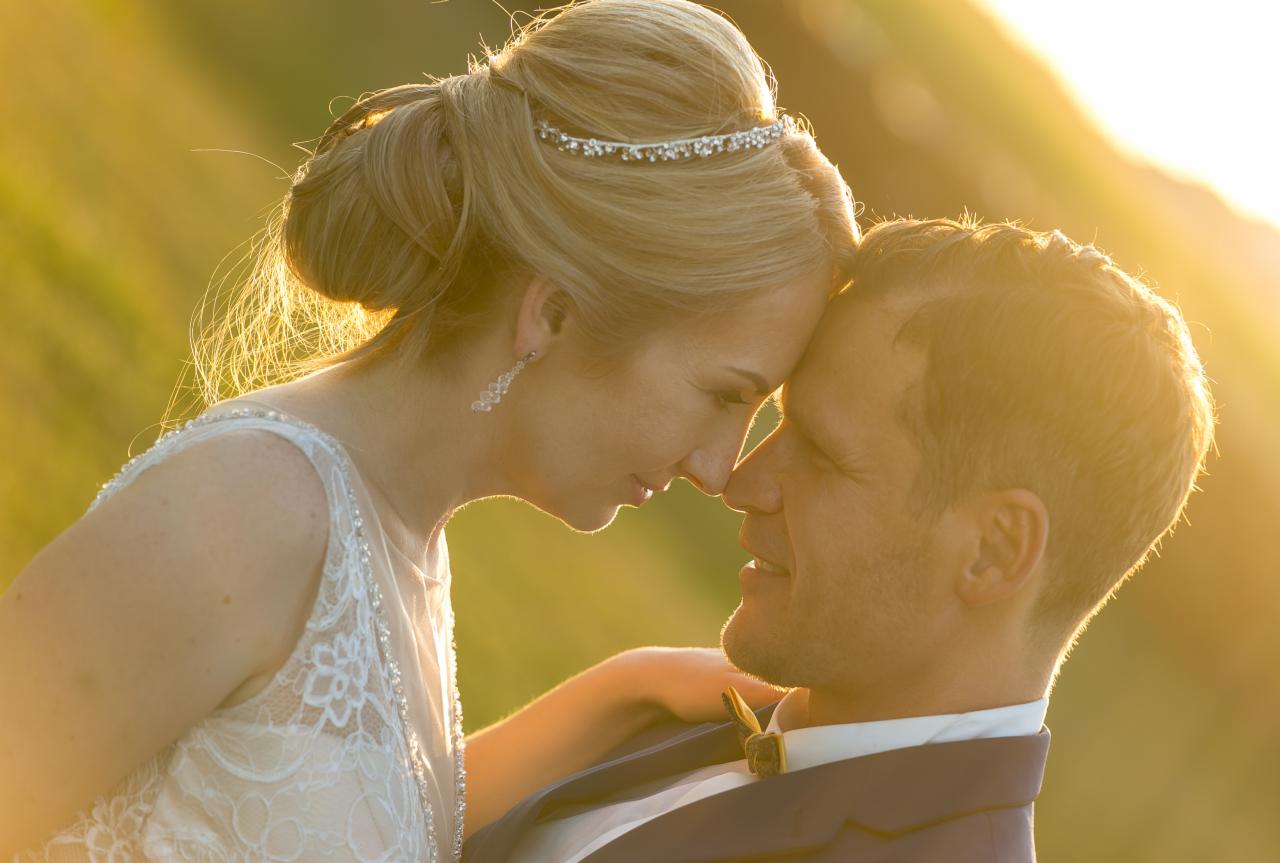 Para młoda w blasku zachodzącego słońca: panna młoda w koronkowej sukni i diademie, pan młody w granatowym garniturze z muszką, dotykają się czołami na tle zieleni.