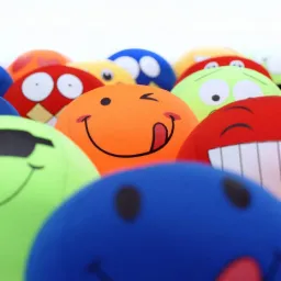 Stos pluszowych, okrągłych poduszek w różnych kolorach z nadrukowanymi emotikonami o różnych wyrazach twarzy, w tym pomarańczowa z wyciągniętym językiem.