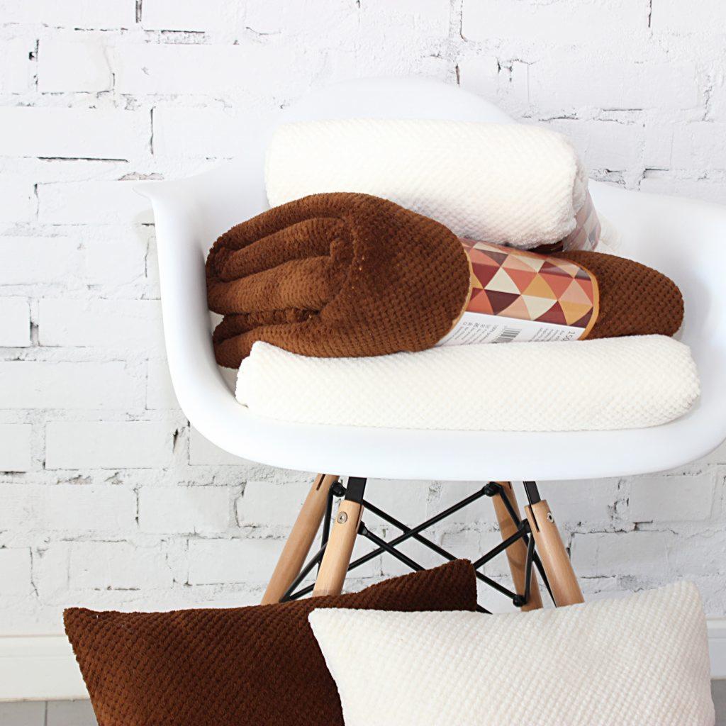 Stos brązowych i kremowych tkanin o fakturze wafelkowej ułożonych na białym krześle na tle ściany z białej cegły, z poduszkami w kolorach tkanin u dołu.