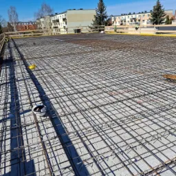 Siatka zbrojeniowa ułożona na betonowej płycie fundamentowej, widoczne kaski ochronne i żółte barierki, w tle budynki mieszkalne pod błękitnym niebem.