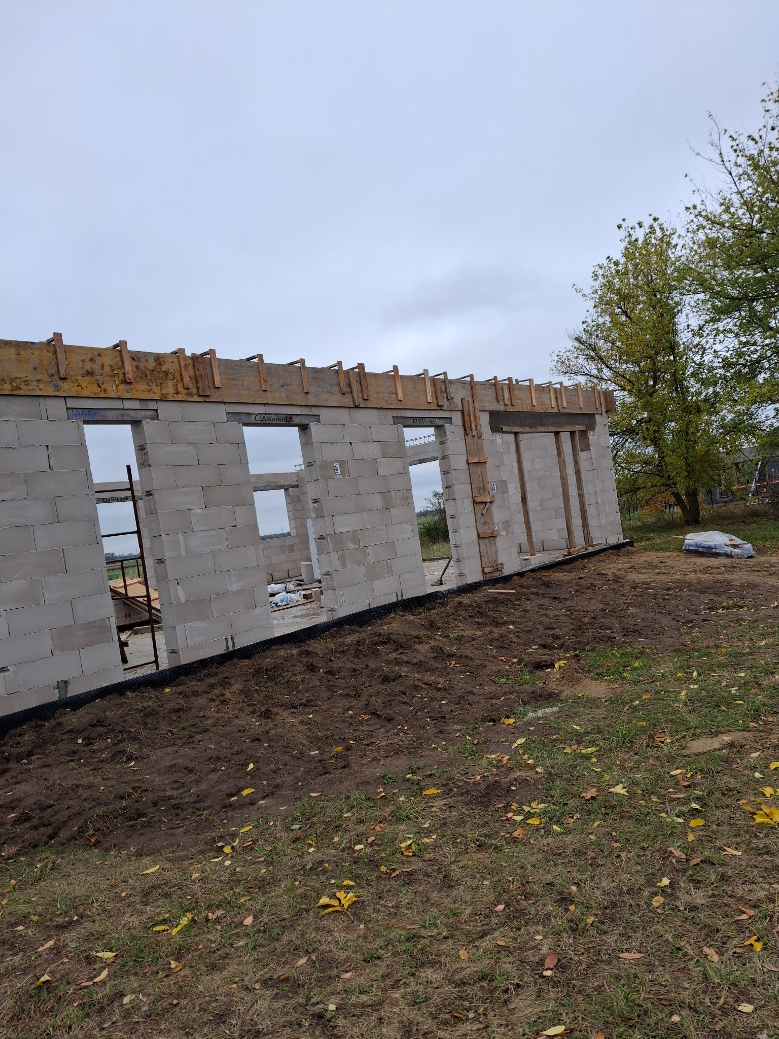 Wznoszone ściany budynku z betonu komórkowego z widocznym szalunkiem nad otworami okiennymi, na zewnątrz teren budowy z workami zaprawy.