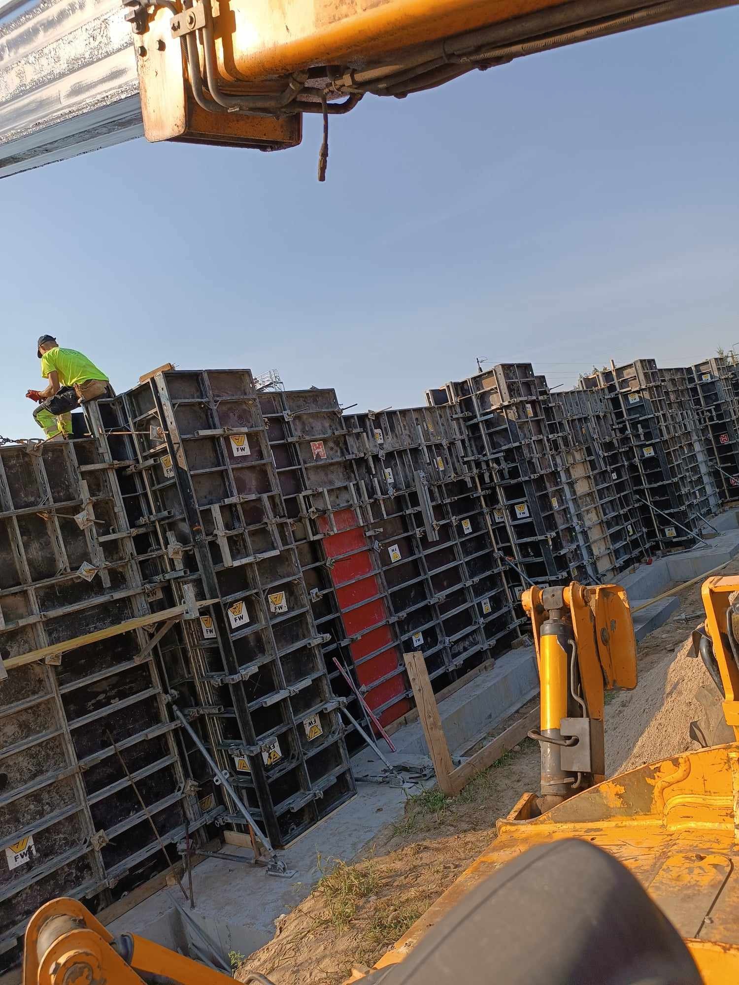 Montaż szalunków systemowych na budowie, widoczny pracownik w odblaskowej kamizelce, elementy dźwigu i żółta maszyna budowlana w tle.