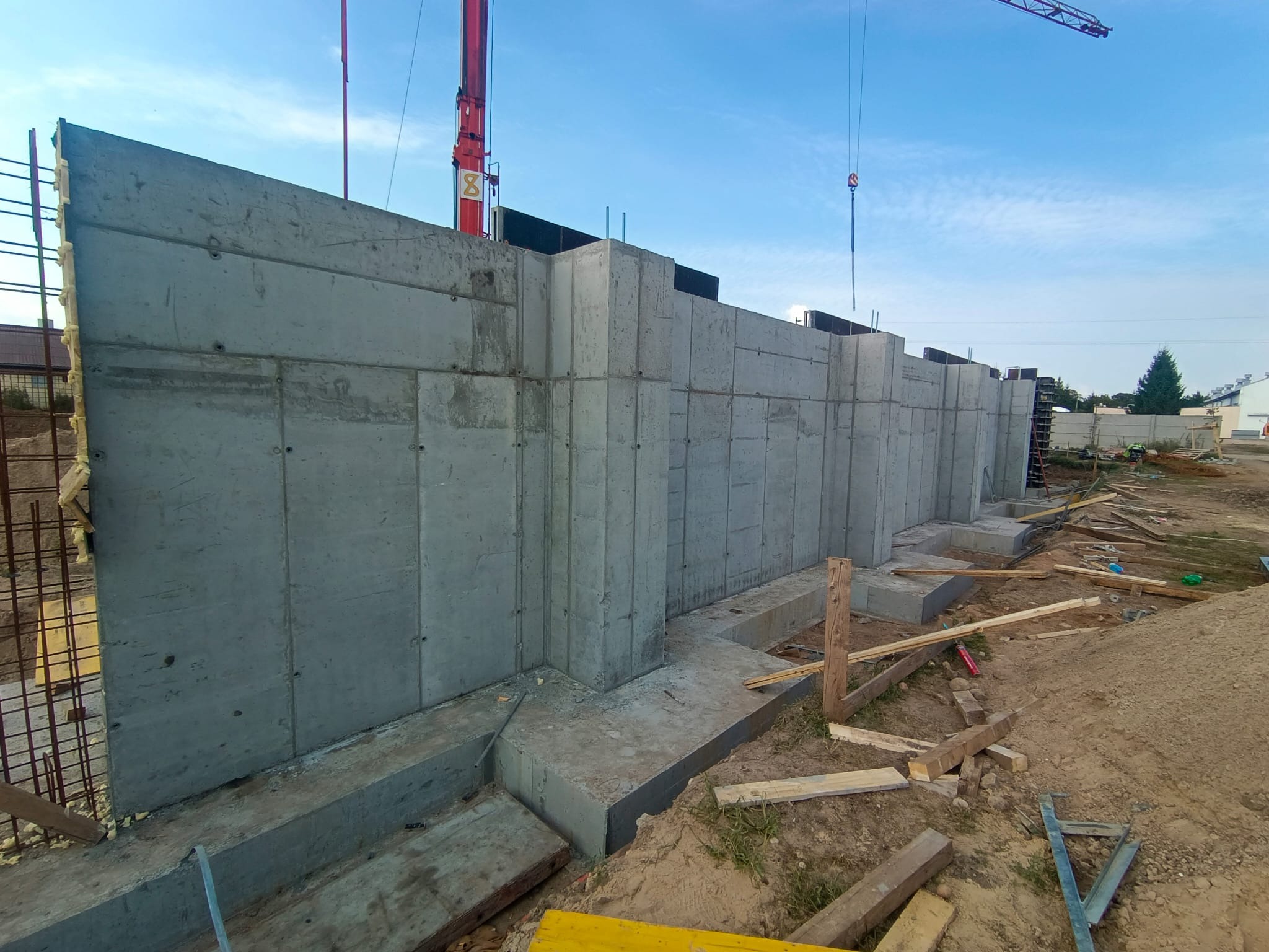 Wylane betonowe ściany fundamentowe z widocznymi śladami szalunków, częściowo zasypane ziemią, z dźwigiem w tle na placu budowy.