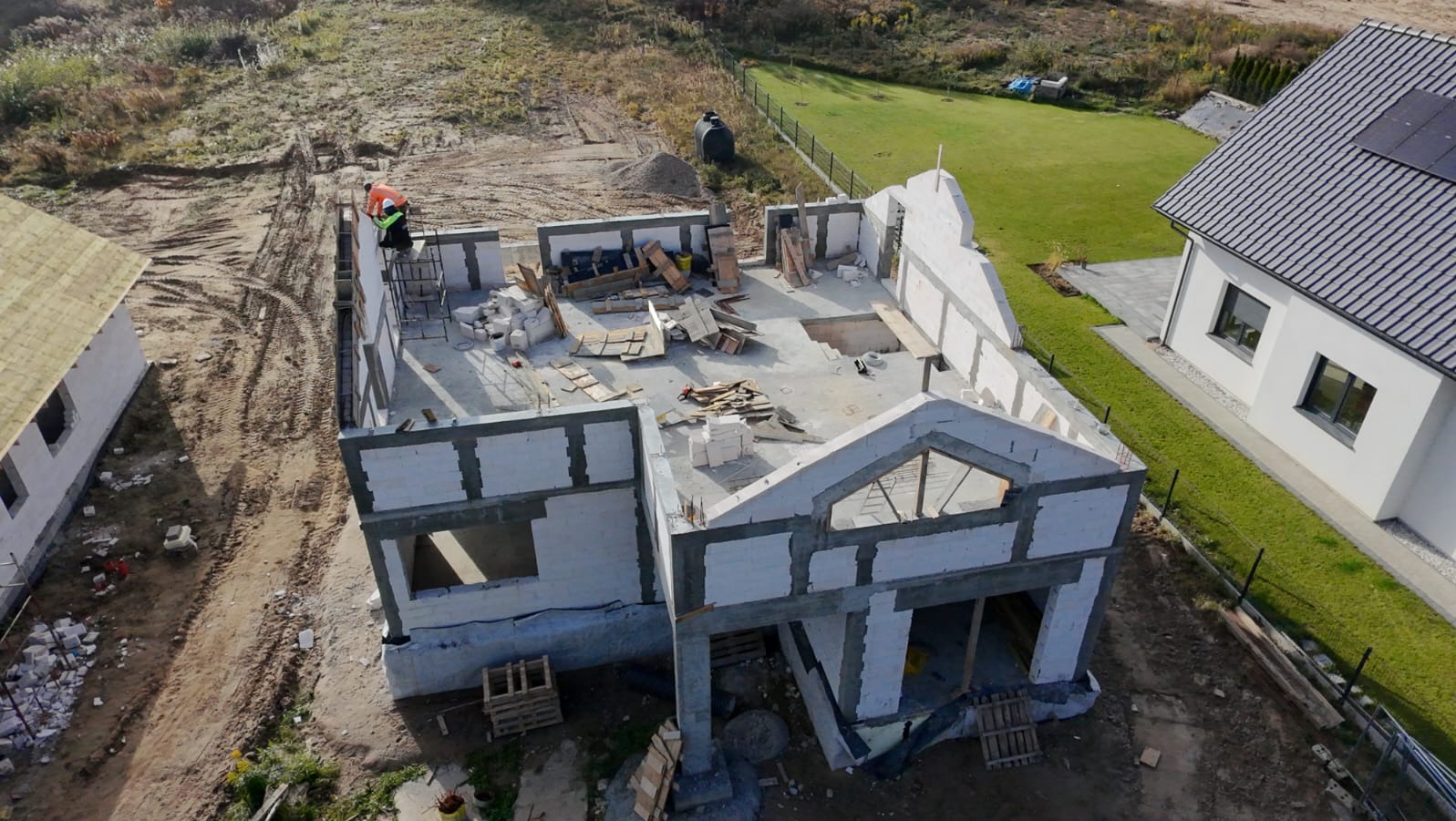 Budowa domu jednorodzinnego w stanie surowym otwartym, widok z góry, z pracownikiem w odblaskowej kamizelce na rusztowaniu.