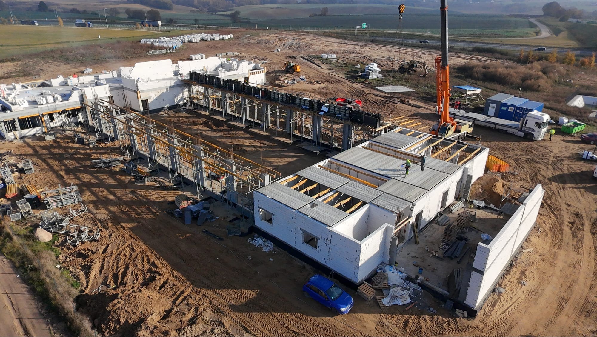 Budowa hali z żelbetu: widok z lotu ptaka na plac budowy z elementami konstrukcyjnymi, dźwigiem i zaparkowanym niebieskim samochodem osobowym