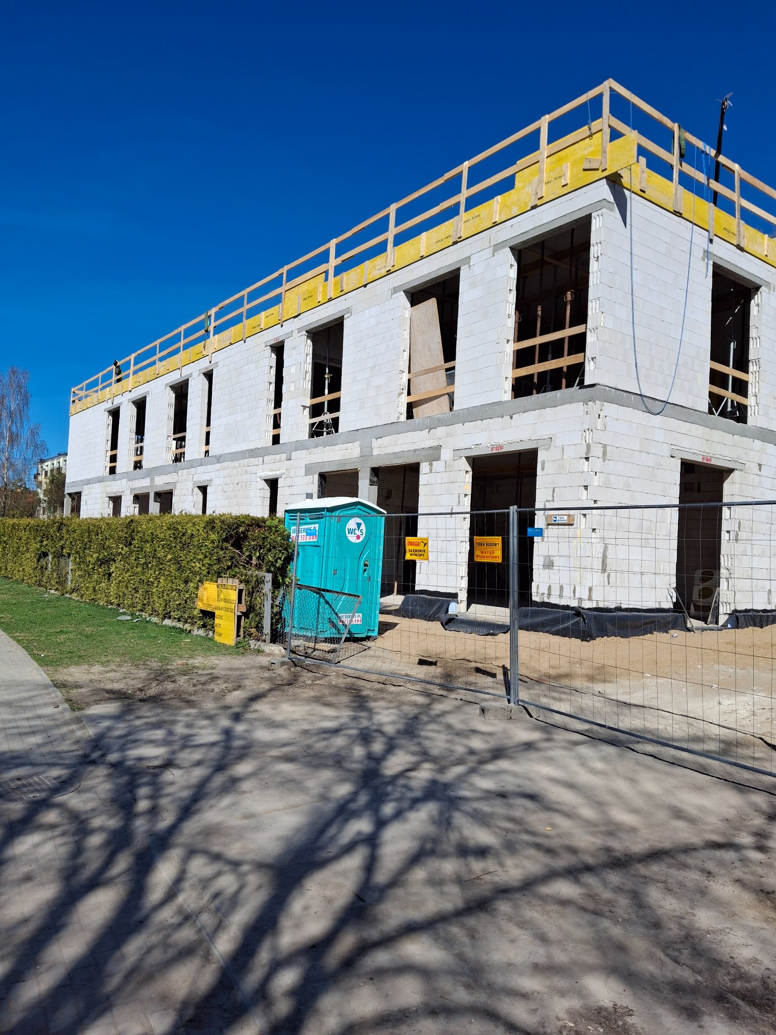 Budynek w stanie surowym otwartym, wzniesiony z białych bloczków, zabezpieczony żółtą barierką na dachu, widoczna przenośna toaleta na placu budowy.