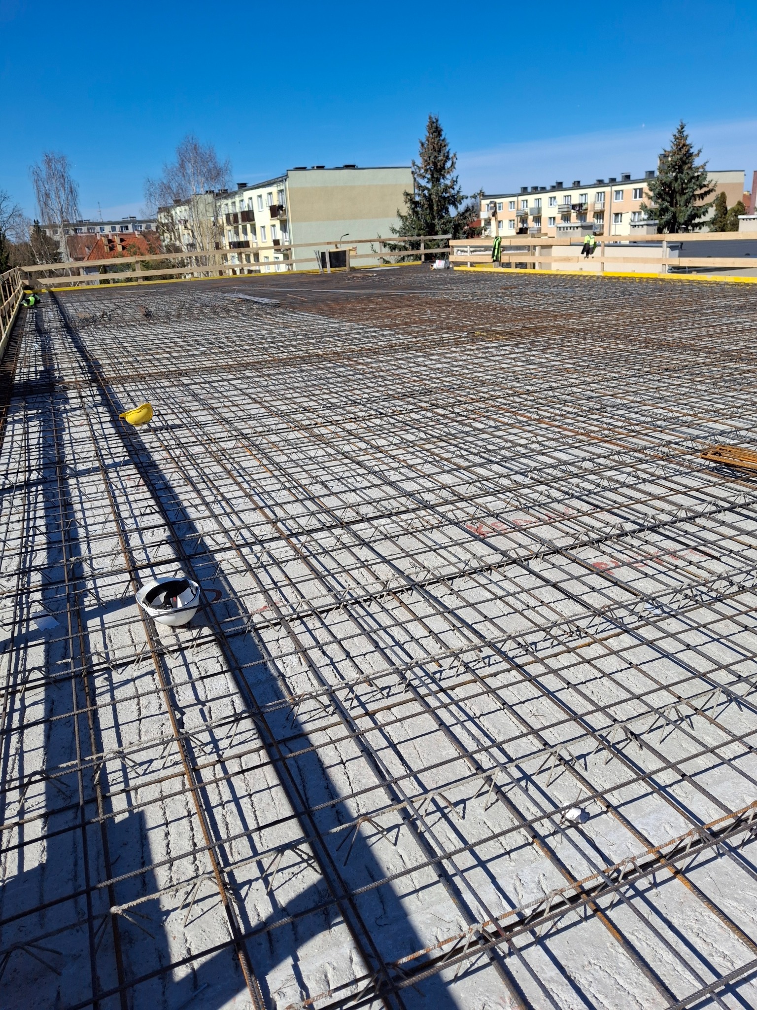Siatka zbrojeniowa ułożona na betonowej płycie fundamentowej, widoczne kaski ochronne i żółte barierki, w tle budynki mieszkalne pod błękitnym niebem.