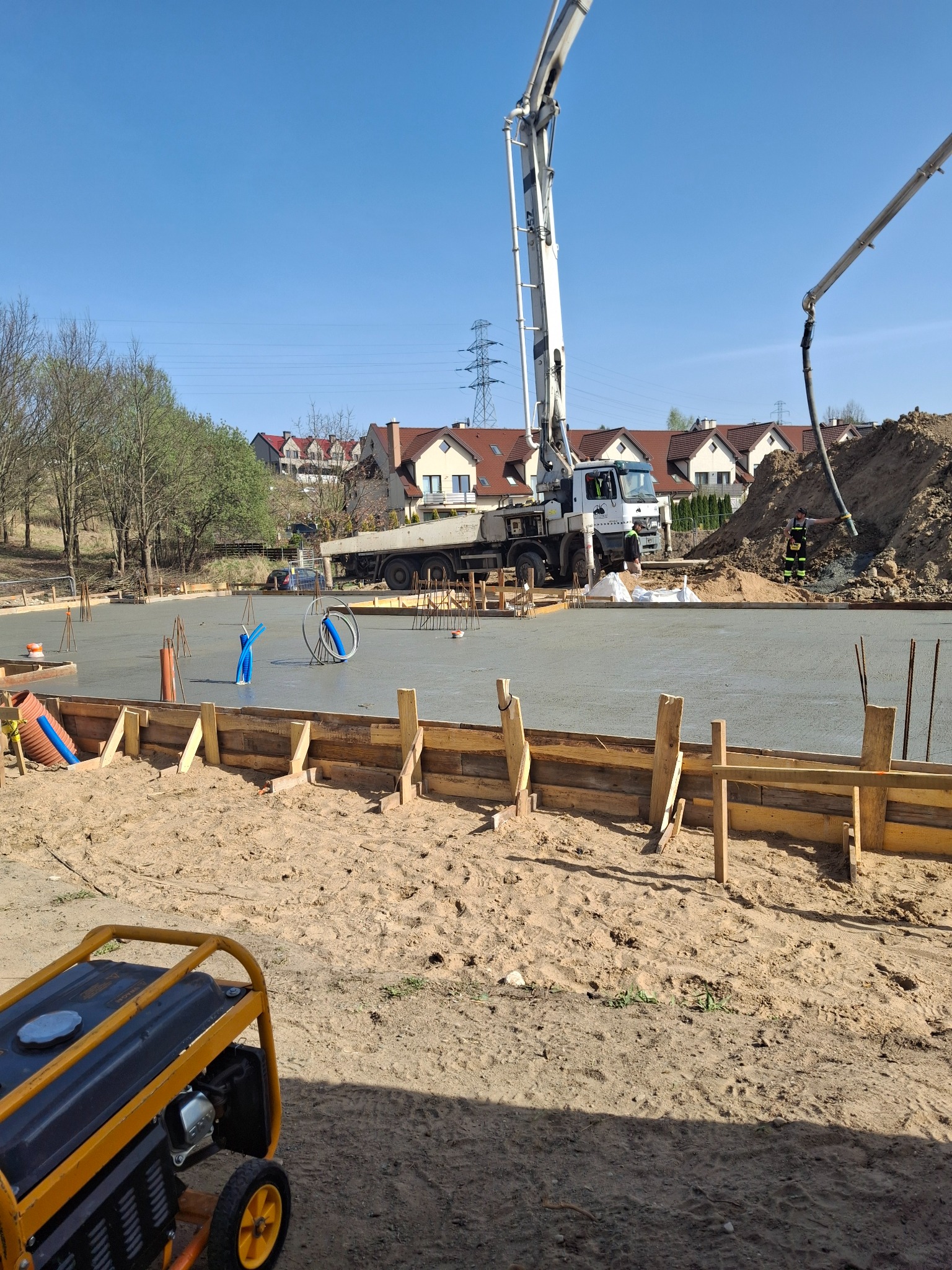 Wylewanie betonu na przygotowany fundament. Widoczny beton, szalunki, pompa do betonu i generator prądu. W tle domy jednorodzinne.