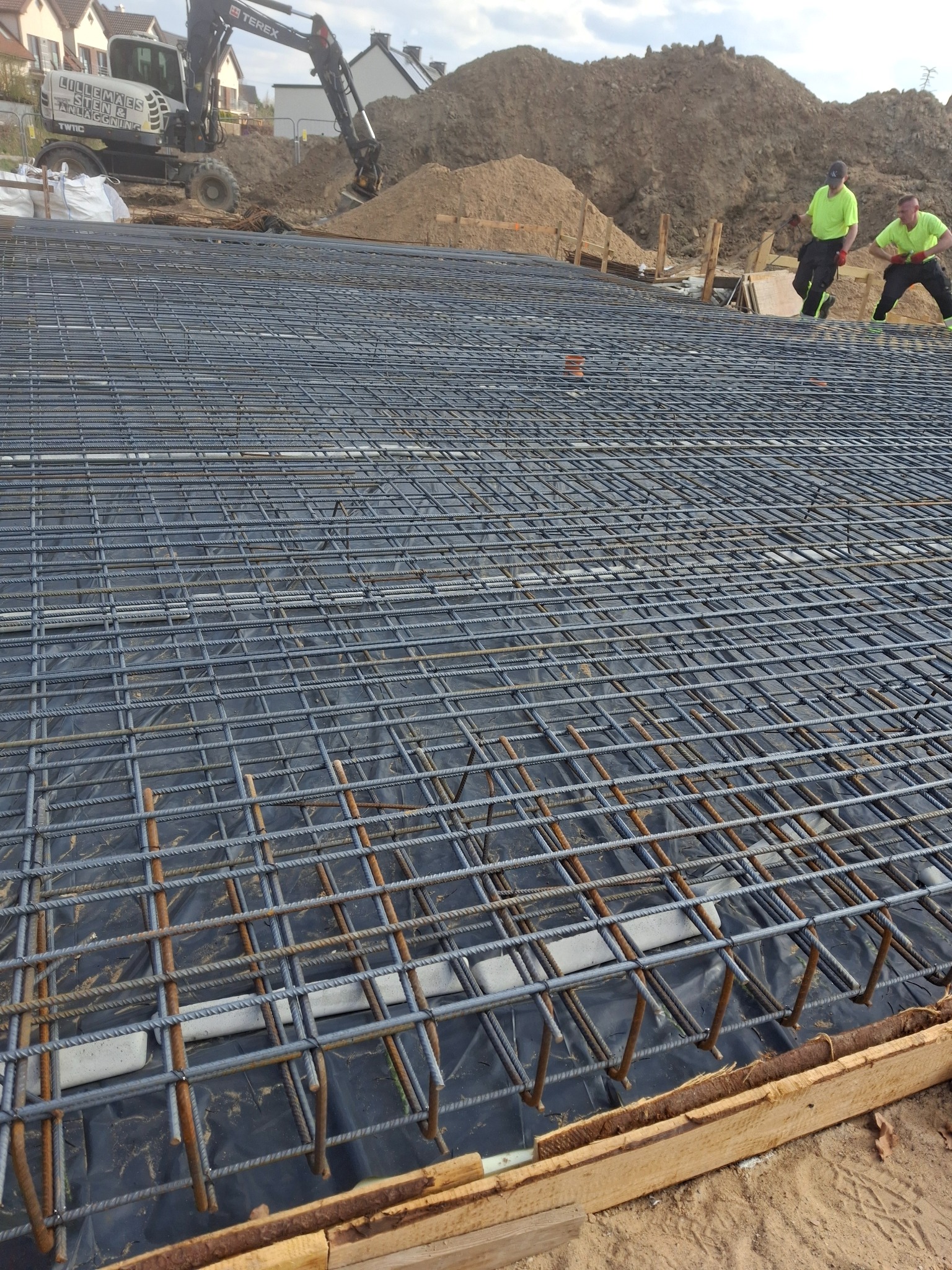 Siatka zbrojeniowa na folii, przygotowana do wylania fundamentów. W tle koparka i dwaj robotnicy w jaskrawych koszulkach. Widok z bliska na konstrukcję.