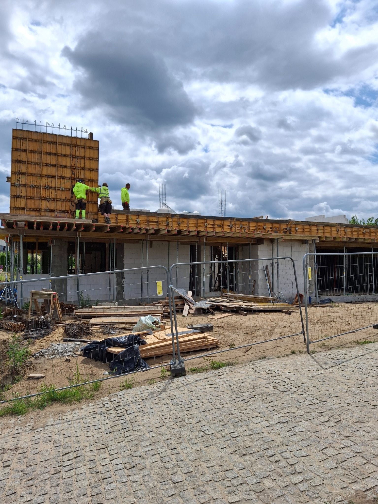 Budowa domu jednorodzinnego: ekipa w żółtych ubraniach pracuje na stropie budynku w trakcie budowy, widoczne szalunki i zbrojenia, ogrodzenie budowy.