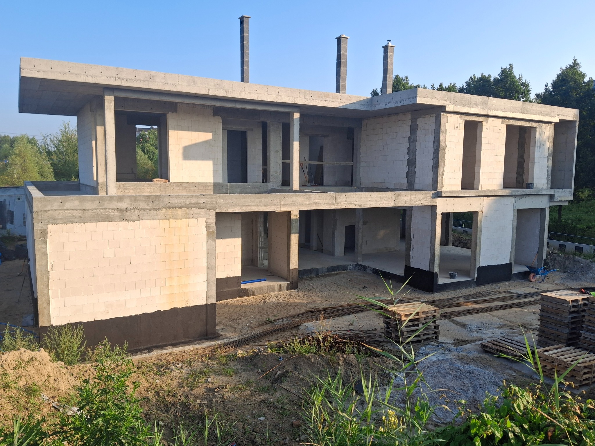 Surowy stan dwukondygnacyjnego domu z betonu komórkowego w trakcie budowy, z widocznymi kominami i izolacją fundamentów, otoczony zielenią.