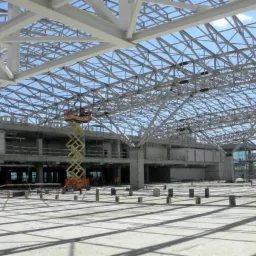 Wnętrze hali w trakcie budowy z widoczną konstrukcją stalową dachu, podnośnikami nożycowymi i betonową posadzką w cieniu.