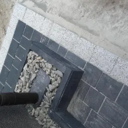 Wykończenie elewacji domu z użyciem szarej kostki brukowej i białego kamienia dekoracyjnego wokół rury spustowej.