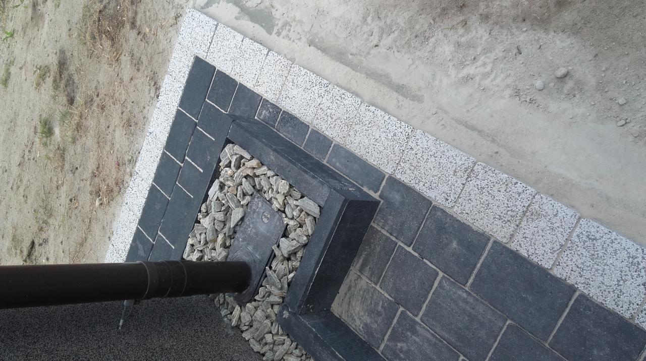 Wykończenie elewacji domu z użyciem szarej kostki brukowej i białego kamienia dekoracyjnego wokół rury spustowej.