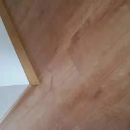 Jasnobrązowe panele podłogowe ułożone przy białej ścianie z widoczną listwą przypodłogową.