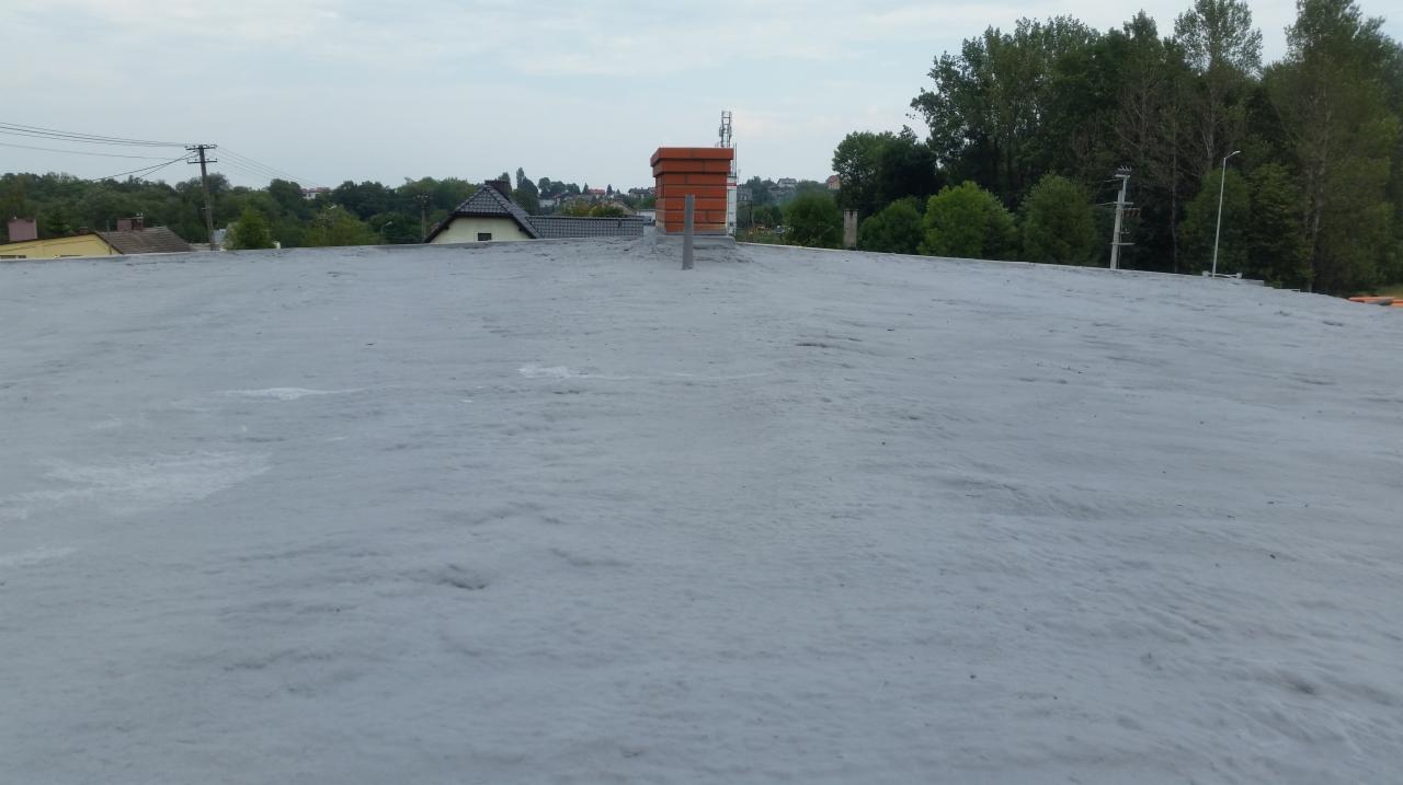 Szare ocieplenie pianką PUR na płaskim dachu budynku, widoczny komin z cegły i otoczenie z drzewami w tle.
