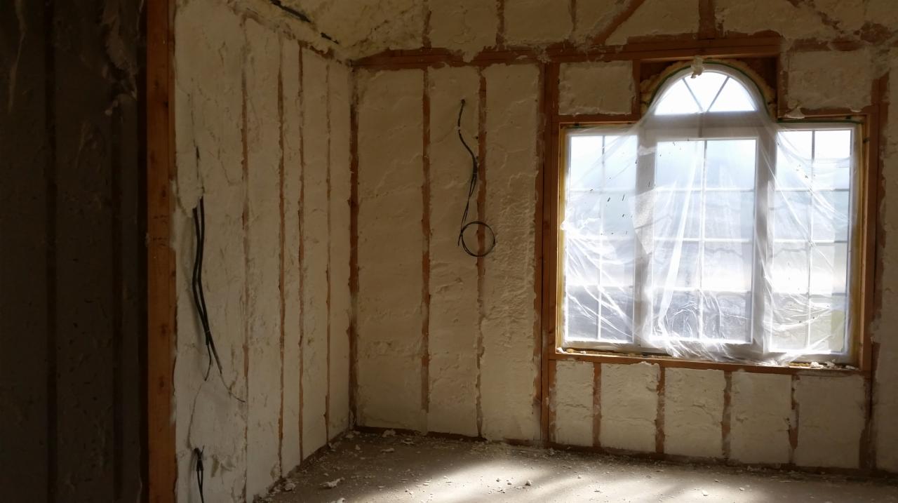 Wnętrze budynku w trakcie ocieplania pianką PUR, widoczne drewniane listwy konstrukcyjne oraz wystające przewody elektryczne, okno zabezpieczone folią.