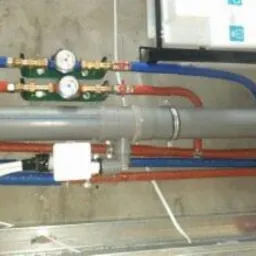 Instalacja wodna i kanalizacyjna z rurami w kolorach czerwonym i niebieskim, widoczne zawory i liczniki, szare rury kanalizacyjne, biała ściana w tle.