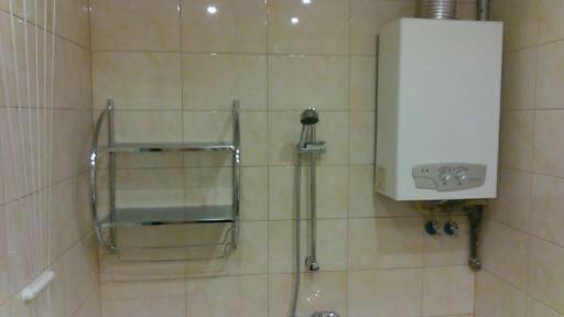 Jasna łazienka z beżowymi płytkami, chromowaną półką i prysznicem oraz białym piecem gazowym z widocznymi pokrętłami.