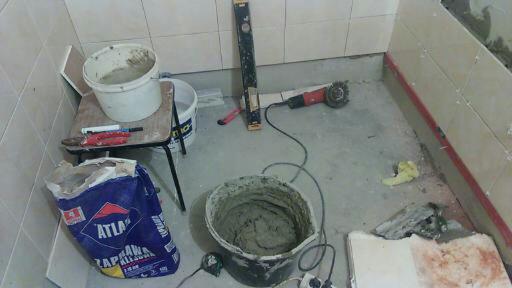 Przygotowania do układania płytek w łazience: wiadro z zaprawą Atlas, poziomica, szlifierka kątowa i częściowo wykończone ściany z jasnych płytek.