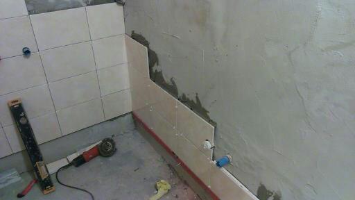 Ściana łazienki w trakcie układania płytek ceramicznych, widoczny poziomica, szlifierka kątowa i fragment surowej ściany z pozostałościami zaprawy.