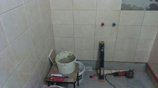 Ściana łazienki w trakcie układania jasnych, kwadratowych płytek; widoczne narzędzia: poziomica, szlifierka kątowa, wiadro z zaprawą i pistolet do fugowania, oraz otwory na baterię.