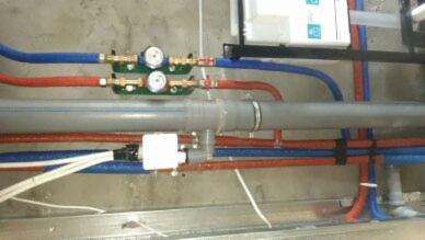 Instalacja wodna i kanalizacyjna z rurami w kolorach czerwonym i niebieskim, widoczne zawory i liczniki, szare rury kanalizacyjne, biała ściana w tle.