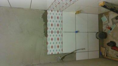 Montaż płytek ściennych w łazience, widoczna zaprawa na ścianie, ułożone płytki z wzorem oraz białe, narzędzia pracy, etap prac wykończeniowych.