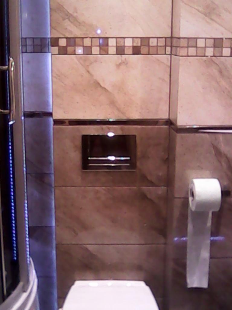 Wykończona łazienka z marmurowymi płytkami, dekoracyjnym pasem mozaiki, podtynkową spłuczką i toaletą, z widocznym fragmentem kabiny prysznicowej z ledowym oświetleniem.