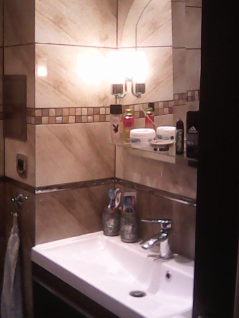 Wykończona łazienka z umywalką, baterią i lustrem z oświetleniem. Beżowe płytki na ścianach z dekoracyjnym pasem mozaiki. Na półce kosmetyki i akcesoria łazienkowe.