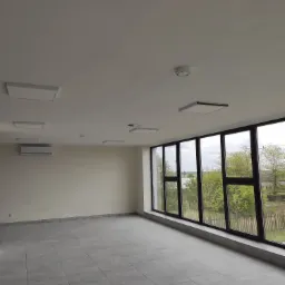 Wnętrze nowo wykończonego pomieszczenia z szarą podłogą, dużymi oknami z widokiem na zieleń i zainstalowanym oświetleniem sufitowym typu LED panel oraz klimatyzacją ścienną.