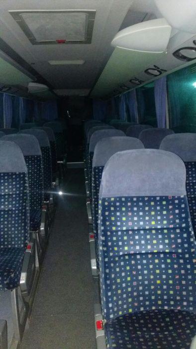 Wnętrze autobusu z rzędami niebieskich foteli w kwadraty, widoczne okna z zasłonkami i oświetlenie sufitowe.