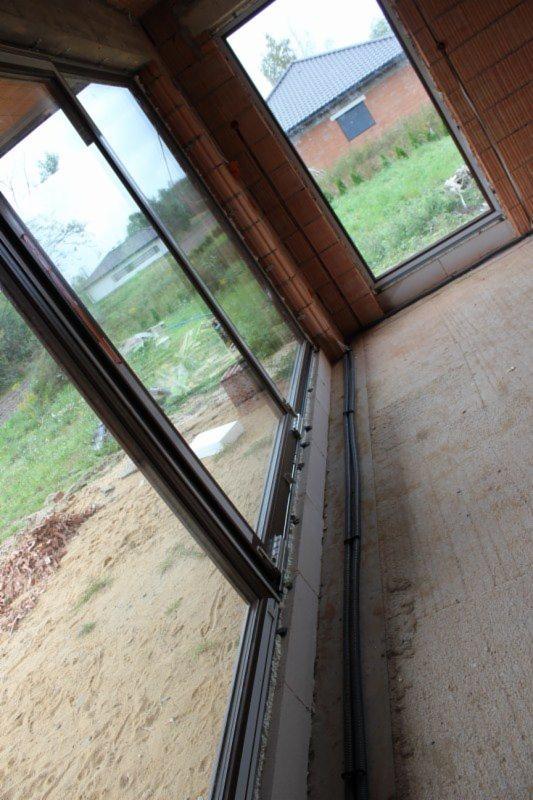 Wnętrze surowego budynku z widocznymi oknami w trakcie montażu, przewody elektryczne w peszlu biegnące wzdłuż ściany.