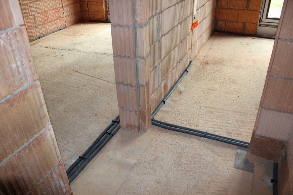 Wnętrze budynku w trakcie budowy z widocznymi przewodami w peszlach, przymocowanymi do podłoża i ścian z czerwonej cegły dziurawki.