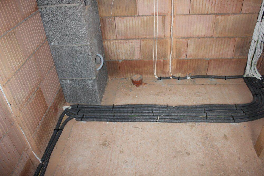 Kable elektryczne w peszlach ułożone wzdłuż ściany z cegły ceramicznej dziurawki i betonowego słupka, widoczne białe przewody i fragment instalacji kanalizacyjnej w podłodze.