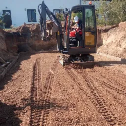 Operator w minikoparce Volvo EC18E wyrównuje teren w wykopie, widoczne ślady gąsienic i drewniane obramowanie.