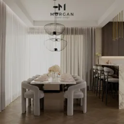 Nowoczesna jadalnia z marmurowym stołem i szarymi krzesłami na tle jasnych zasłon. Nad stołem wiszą designerskie lampy. Widoczny fragment blatu kuchennego z barkiem.