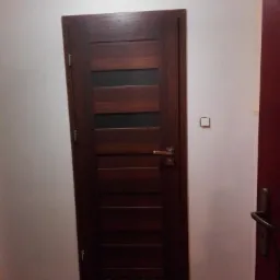 Ciemne, drewniane drzwi wewnętrzne z poziomymi przeszkleniami, zamontowane w białej ścianie, widoczne na tle podłogi imitującej drewno.