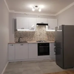 Nowoczesna kuchnia z białymi szafkami, mozaikową ścianą nad blatem, czarną armaturą i srebrną lodówką, oświetlona lampami sufitowymi i listwami LED pod szafkami.