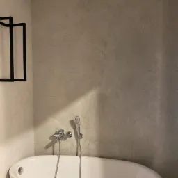 Nowoczesna łazienka z owalną białą wanną i ścianą wykończoną dekoracyjnym tynkiem w odcieniach szarości, z minimalistyczną czarną półką na ścianie.