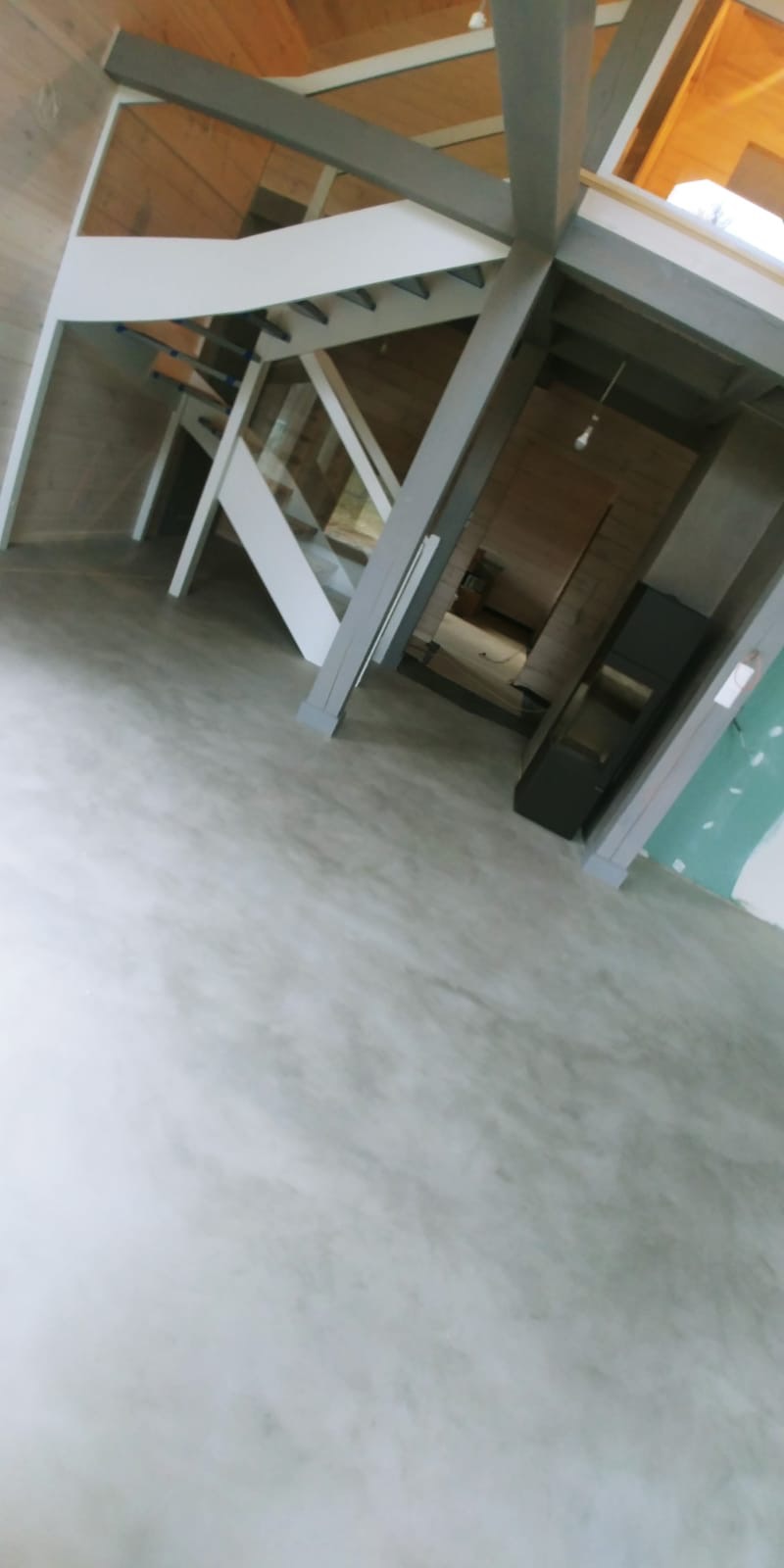 Jasnoszara, jednolita podłoga w nowoczesnym wnętrzu z drewnianymi elementami konstrukcyjnymi i schodami; widok na dalsze pomieszczenia.