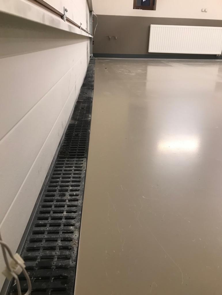 Beżowa, błyszcząca podłoga z odpływem liniowym przy ścianie garażu, widoczny fragment grzejnika i okna w tle.