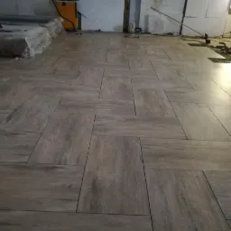 Ułożona podłoga z płytek imitujących drewno w jodełkę, widoczne narzędzia i wiadro z klejem w tle.