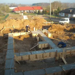Wylany fundament pod budynek mieszkalny, widoczne drewniane szalunki i dwóch robotników w trakcie prac, w tle zabudowa jednorodzinna i samochód dostawczy.