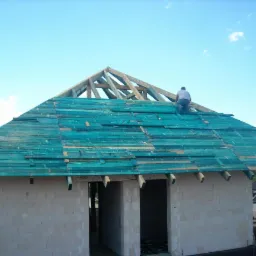 Budowa domu jednorodzinnego: murarz układa deski na dachu, w tle widoczny biały bus i błękitne niebo.