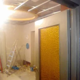 Wnętrze w trakcie remontu: widoczny stelaż sufitu podwieszanego z okrągłym elementem dekoracyjnym, drabina, ściany przygotowane do malowania oraz drzwi z pomarańczową szybą o fakturze plastra miodu.