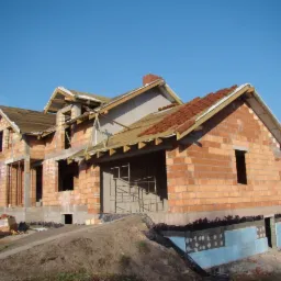 Niedokończony dom z czerwonej cegły Porotherm, z widoczną izolacją fundament&oacute;w i częściowo ułożonym dachem, na tle błękitnego nieba.