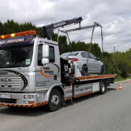 Srebrna laweta MAN z podnośnikiem holuje srebrny samochód osobowy na drodze z pomarańczowymi pachołkami, w tle zielone wzgórza i budynki.