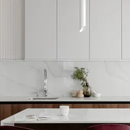 Nowoczesna kuchnia z białymi szafkami, marmurowym blatem i backsplashiem, oświetleniem tubowym oraz elementami drewnopodobnymi, z wyspą, na której leży otwarta książka i stoi biały kubek.