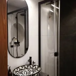 Nowoczesna łazienka z owalnym lustrem w czarnej ramie, umywalką ceramiczną w czarno-białe wzory i kabiną prysznicową z przesuwanymi drzwiami.